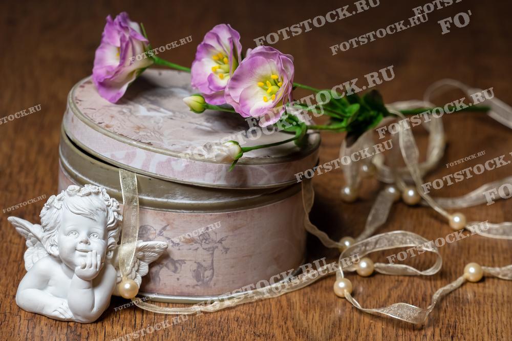 На фотографии изображены светло-фиолетовые цветы эустомы, расположенные на винтажной металлической шкатулке с рисунком. Рядом видна статуэтка ангела и декоративная лента с жемчужинами, что придает композиции нежность и романтическое настроение. 
Это изображение подходит для использования в цветочных бутиках, для создания атмосферы на праздновании Дня Святого Валентина или свадьбы, а также как элемент декора для дома.